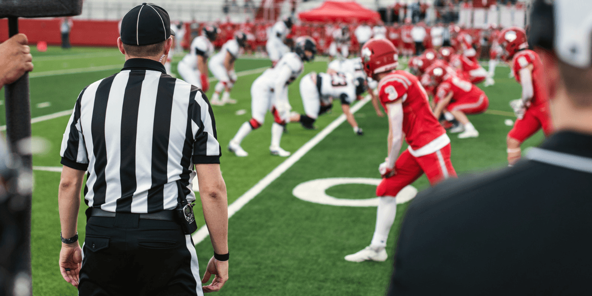A referee observes two football teams lined up on the field ready for play